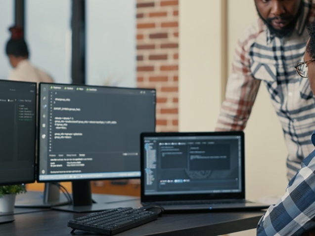 Focused software developer writing code on laptop looking at multiple screens with programming language is interrupted by colleague coder asking for advice. Programmers doing online cloud computing.