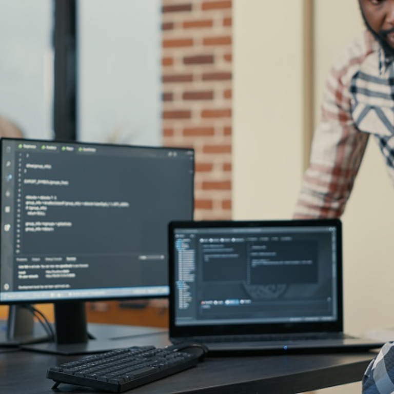 Focused software developer writing code on laptop looking at multiple screens with programming language is interrupted by colleague coder asking for advice. Programmers doing online cloud computing.