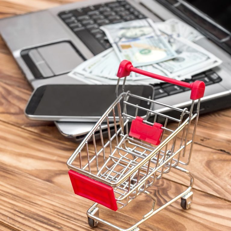 Shopping cart and laptop with money and smartphone on the table. Online shopping and Ecommerce.